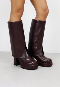 Botas de cuero burdeos, brillantes, hasta la rodilla, con diseño de abertura lateral, suelas de plataforma gruesas y tacones bloque robustos. Textura suave en general.