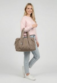 Woman in pink sweater, light jeans, and white sneakers holding a large taupe tote bag with a round silver tag.