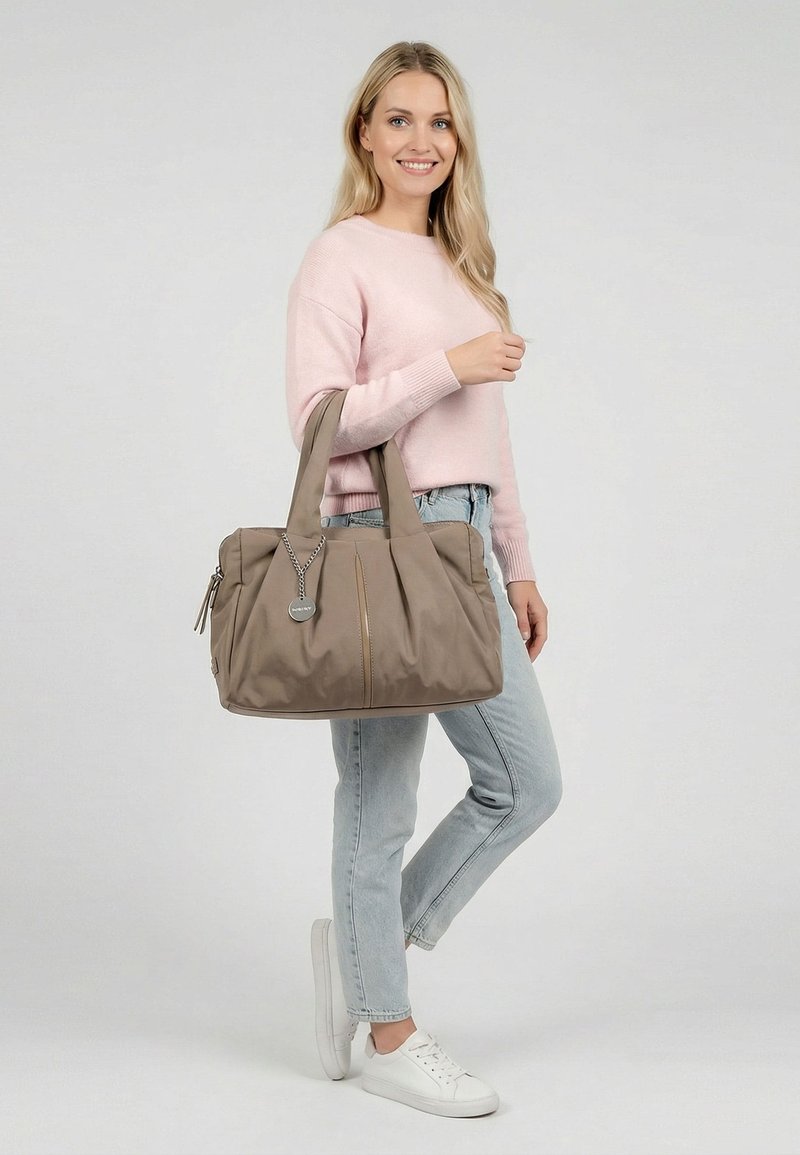 Woman in pink sweater, light jeans, and white sneakers holding a large taupe tote bag with a round silver tag.