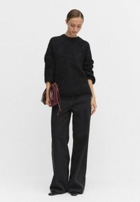 Svart stickad tröja med lösa ärmar, parat med vida svarta byxor med subtilt prickmönster. Håller en mörkt vinröd väska.