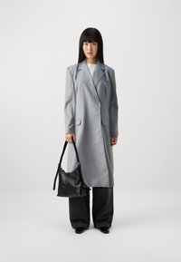 Grå lang frakk med kneppede slag, to frontlommer, båret over en lys topp, kombinert med mørke vide jeans og en svart håndveske.