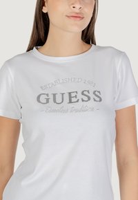 T-shirt bianco in cotone con maniche corte e scollatura rotonda, caratterizzato da un logo "GUESS" in rilievo argentato e dal testo "Established 1981" sopra.