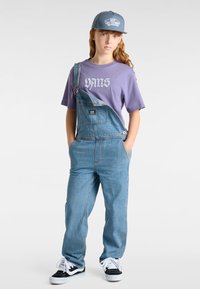 Los overoles de mezclilla presentan tirantes ajustables y un bolsillo frontal, combinados con una camiseta gráfica de color púrpura claro y zapatillas en blanco y negro.