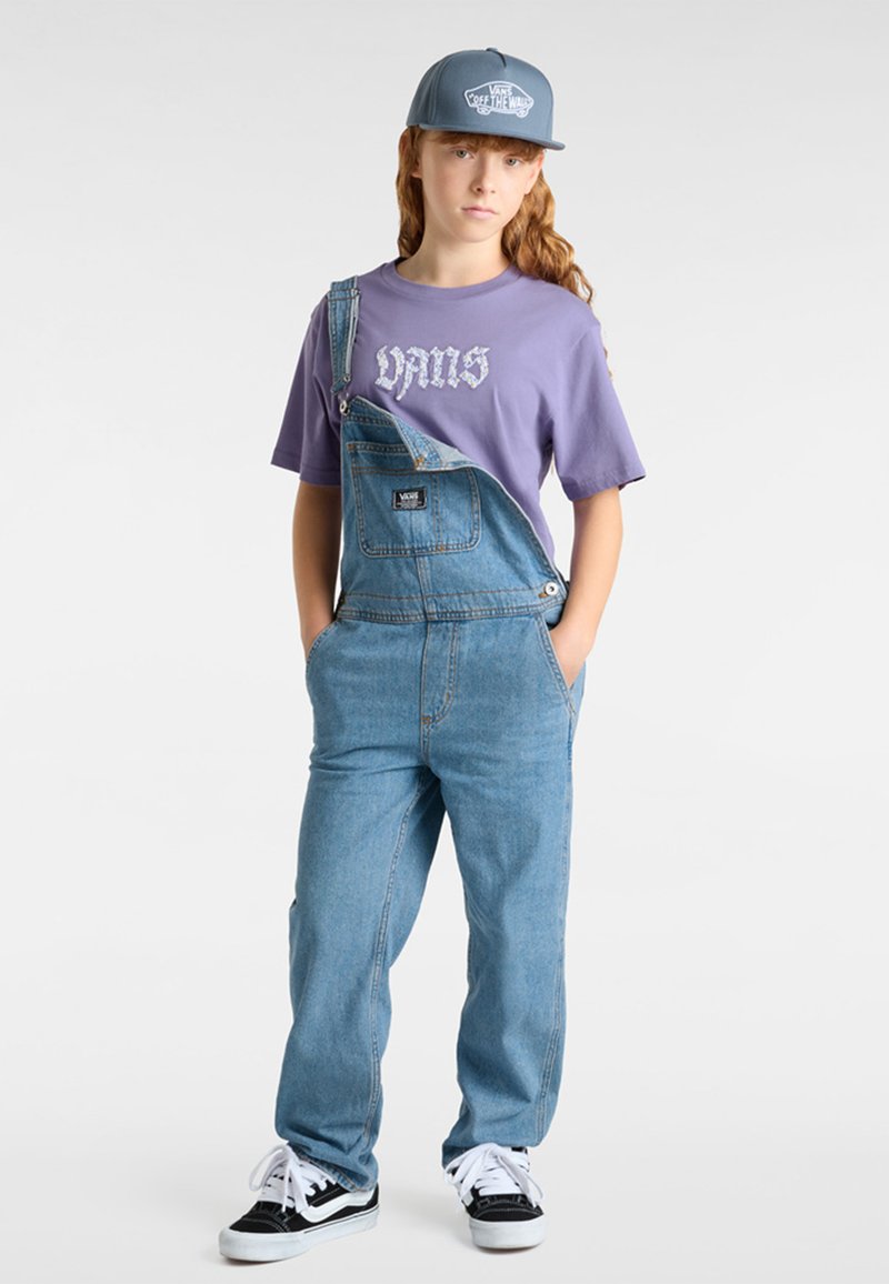 Los overoles de mezclilla presentan tirantes ajustables y un bolsillo frontal, combinados con una camiseta gráfica de color púrpura claro y zapatillas en blanco y negro.