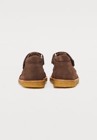 Chaussures en daim marron avec un petit talon, de forme arrondie et avec des ouvertures à l'arrière. Elles présentent une fermeture à sangle unique avec un accent doré.