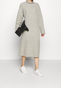 Robe tricotée côtelée gris clair avec un col haut et des manches longues, associée à des baskets blanches et tenant des bottes à talons noires.