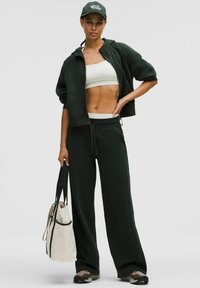 Tmavozelená mikina s kapucňou a zodpovedajúce tepláky vyrobené z mäkkého materiálu. Modelka nesie svetlú tašku a má na sebe priliehavý krémový crop top a čiapku.