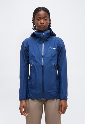 Nő kék zipzáras kapucnis dzsekiben és bézs nadrágban, egyszínű fehér háttér előtt áll.