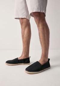 Black fabric slip-on shoes with a woven texture, featuring a natural rope sole and a small rear loop for easy wear.