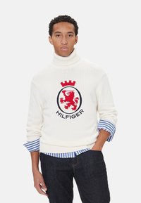 Jersey de cuello alto blanco con emblema de león rojo, texto "HILFIGER" y borde azul marino. Puños a rayas azul y blanca visibles en las mangas.