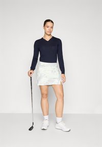 Tengerészeti kék, hosszú ujjú V-kivágású felső, világoszöld mintás golf szoknyával párosítva. A modell golfütőt tart, és fehér sportcipőt visel.