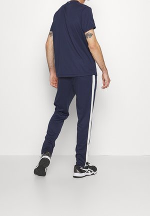 Homme portant un t-shirt bleu marine et un pantalon de survêtement assorti avec des bandes blanches sur les côtés, chaussures de sport noires et blanches, debout contre un mur blanc.