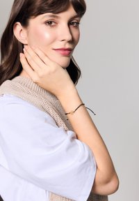 Czarny sznurkowy bransoletka z złotym kwiatowym zawieszką na nadgarstku. Model nosi beżowy sweter i białą koszulę z krótkim rękawem. Neutralne tło.