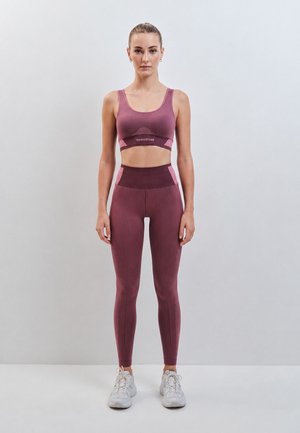 Donna in piedi di fronte, indossa reggiseno sportivo e leggings abbinati color malva e rosa con scarpe da ginnastica bianche su uno sfondo chiaro e uniforme.