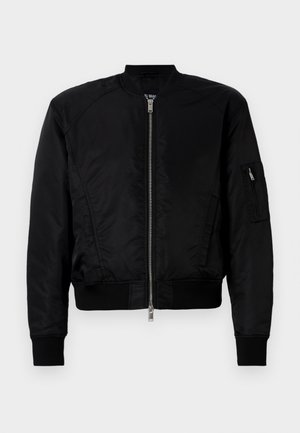 Mies pukeutuneena mustaan bomber-takkiin, mustaan graafiseen T-paitaan, vaalean sinisiin leveälahkeisiin farkkuihin ja mustiin kenkiin yksiväristä valkoista taustaa vasten.