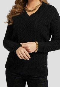 Pull en tricot noir avec un col en V et un motif en câble texturé. Équipé de poignets et d'un ourlet côtelés, associé à un bracelet en chaîne dorée.