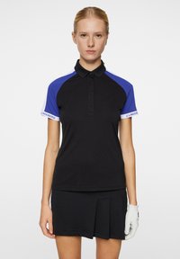 Svart polo skjorta med blå och vita accenter, med en ribbad krage, knäppning fram och logga på ärmen. Kombinerad med en svart plisserad kjol.