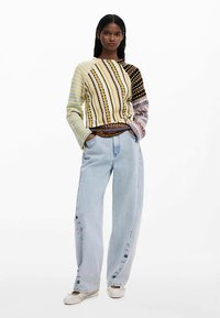 Meer gekleurde gebreide trui met gestreept ontwerp, gedragen met lichtblauwe losse jeans met knoopdetails aan de zijkant en witte canvas schoenen.