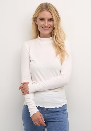 Maglia bianca a maniche lunghe con scollatura alta e orlo ondulato, abbinata a jeans in denim blu. Il tessuto sembra morbido e aderente.