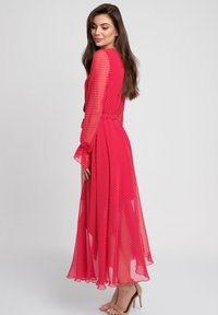 Czerwona maxi sukienka w kropki z przezroczystymi długimi rękawami, dopasowaną talią i zwiewnym dołem. Zawiera falbany na końcach rękawów.