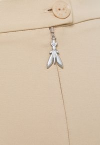 Beige rok stof met een gladde structuur, voorzien van een metalen ritsen trekker in de vorm van een insect en een ronde beige knop aan de bovenkant.