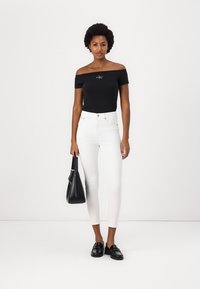 Zwarte off-the-shoulder top met logo, witte skinny jeans, zwarte loafers en een zwarte handtas. Gladde stof en getailleerde silhouet.