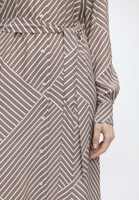 Gros plan d'une robe à rayures marron et blanche boutonnée avec une ceinture à la taille et une main reposant à côté de la hanche.