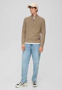 Beiger Reißverschluss-Pullover aus strukturiertem Stoff, kombiniert mit hellblauen Jeans und schwarz-weißen Sneakers. Das Modell trägt eine beigefarbene Mütze.