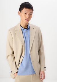 Beige szabott blézer világoskék pólóing felett. A blézer szabott hajtókákkal és fekete gombokkal rendelkezik. Simább anyag, strukturált dizájn és testhezálló fazon.
