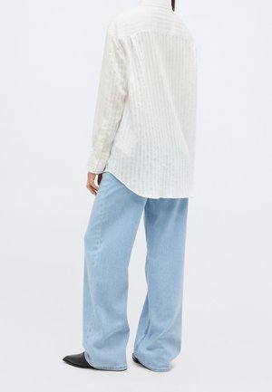 Persoon draagt een los wit shirt met lange mouwen en lichtblauwe wijde jeans, met de rug naar de camera, met zwarte puntige schoenen.