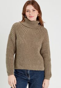 Beige strikket turtle neck trøje med ribbet tekstur, der har et kraftigt design og en let løs pasform. Fremhævet med en høj, foldet krave.