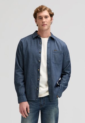 Mladić u plavoj košulji s prugama preko bijele majice i plavih traperica, stoji uz jednobojnu sivu pozadinu.