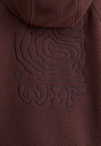 Prenda con capucha de forro polar marrón que presenta un diseño espiral bordado en negro y el logo "WIP", con una textura suave y un ajuste relajado.