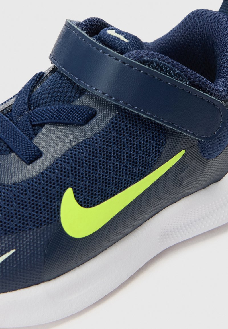 Scarpa sportiva in rete di colore blu navy con cinturino in velcro, dettagli in verde neon e suola bianca. Presenta un tessuto testurizzato e un design curvato.