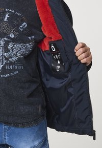 Námornícky kabát s červeným umelým kožúškom vo vnútri, zapínanie na zips a vnútorná etiketa. Nosený cez tmavý tričko s grafikou a džínsy.