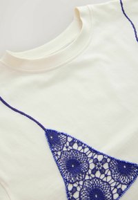 Chemise de couleur crème avec un col rond présentant un motif de dentelle au crochet bleu en forme triangulaire sur le devant.