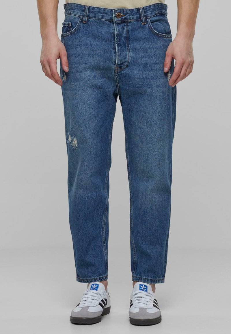 Vaqueros de denim azul claro con un corte relajado, que presentan un detalle desgastado en la rodilla, cinco bolsillos y un cierre de botón. Combinados con zapatillas blancas y grises.
