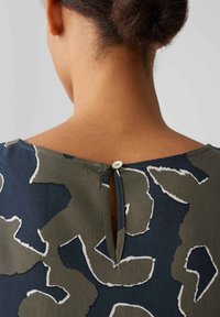 Dark blue blouse featuring an abstract olive green and white pattern with a keyhole opening at the back, finished with a button closure.
