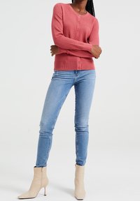 Růžový texturovaný kardigan na knoflíky, kombinovaný s světle modrými úzkými džíny a krémovými kotníkovými botami. Hladká látka a přiléhavý střih.