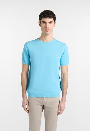 Mladý muž nosiaci svetlomodrú košeľu s krátkym rukávom a malým bielym logom na hrudi a béžové nohavice, stojaci pred jednoliatym bielym pozadím.