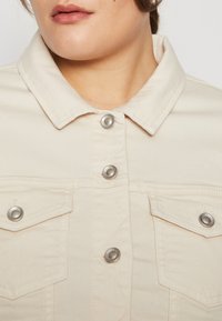 Beige Jeansjacke mit spitzem Kragen, silbernen Druckknöpfen und Brusttaschen mit Steppnähten. Glatte Textur, lässiges Design.