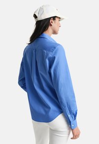 Camicia a maniche lunghe blu in cotone, con colletto button-down, piega sul retro e polsini con bottone, abbinata a pantaloni bianchi e un cappellino.