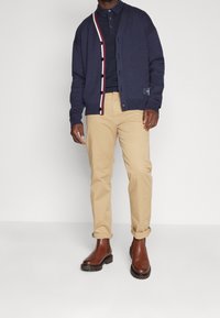 Marineblaues Strickjacke mit rot-weißen Streifendetails, darüber ein marineblaues Poloshirt, kombiniert mit hellbeigen Hosen und braunen Chelsea-Boots mit Knöchelhöhe.