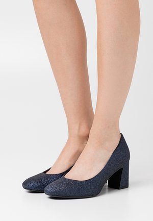 Femme portant des escarpins à talons bloc pailletés bleu marine, se tenant sur une surface blanche unie.