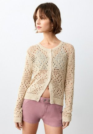 Młoda kobieta patrząca w lewo, ubrana w beżowy rozpinany sweter ażurowy oraz wysokiej talii szorty w kolorze mauve, stojąca na tle jednolitego białego tła.