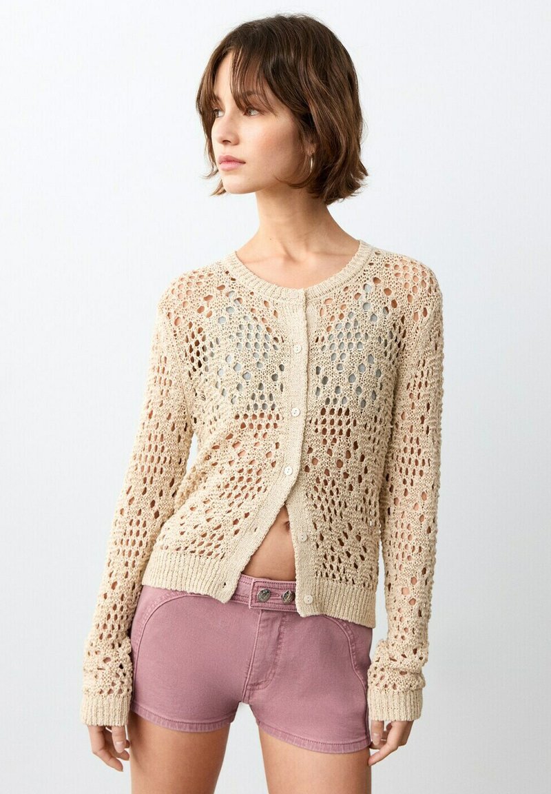 Giovane donna che guarda a sinistra, indossando un cardigan beige a maglia aperta con bottoni e pantaloncini viola a vita alta, in piedi davanti a uno sfondo bianco semplice.