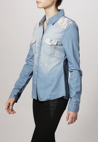Chemise en denim bleu clair avec des accents en dentelle florale sur les épaules et les poches poitrine, avec une fermeture à boutons et des manches ajustées.