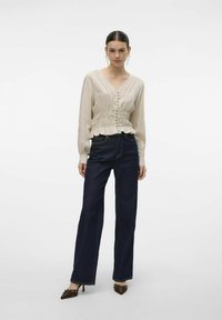 Beige blouse met lange mouwen, een V-hals, een gerimpelde zoom en knoopsluiting, gecombineerd met donkerblauwe high-waisted jeans en bruine hakken.
