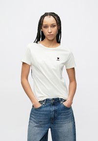 T-shirt creme feita de algodão macio, com um pequeno coração bordô e o texto "Bigger" no lado esquerdo do peito, combinada com calças de ganga azuis.