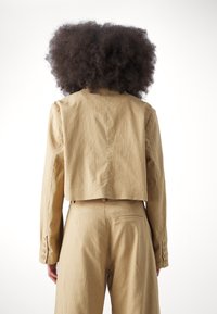 Veste courte beige avec une finition texturée, présentant une coupe droite et des poignets à boutons, assortie à un pantalon taille haute.